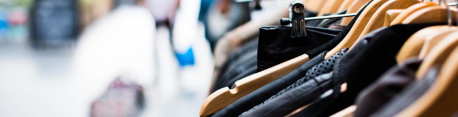 A rack of clothing on wooden hangers in a fashion retail store, with a shopper browsing in the background.