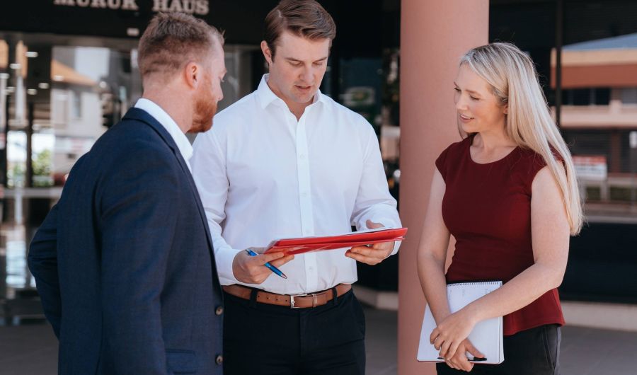 Three people in business attire reviewing documents in a red folder outside a commercial building.
