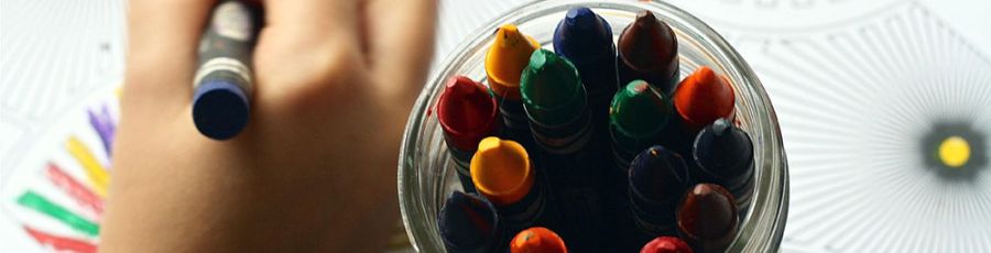 A child's hand colouring a worksheet next to a glass jar of coloured crayons on a classroom table.