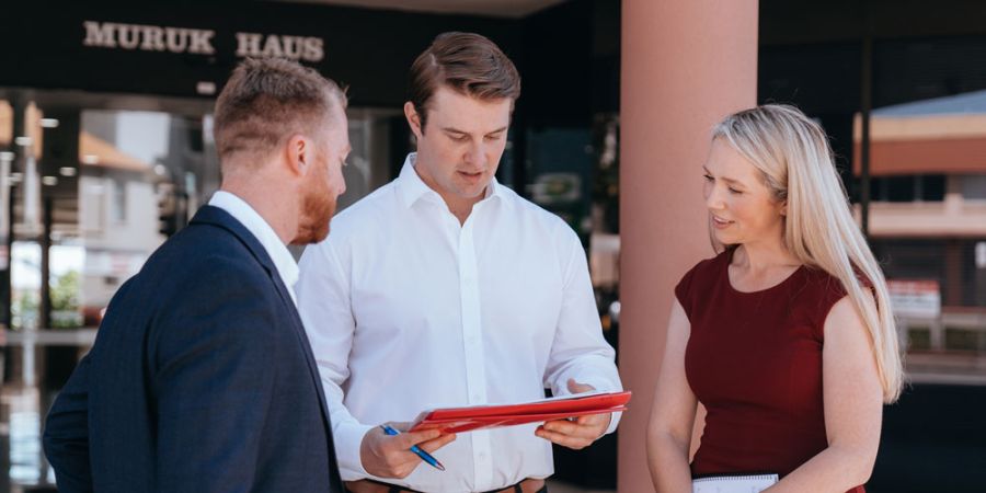 Two men and a woman in business attire discuss documents outside an office building.