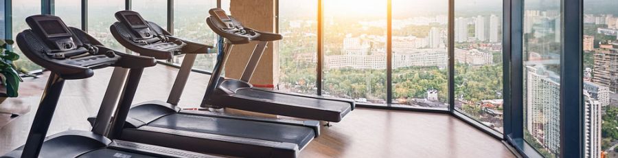 A row of treadmills on a timber floor beside floor-to-ceiling windows overlooking a city skyline at sunrise.