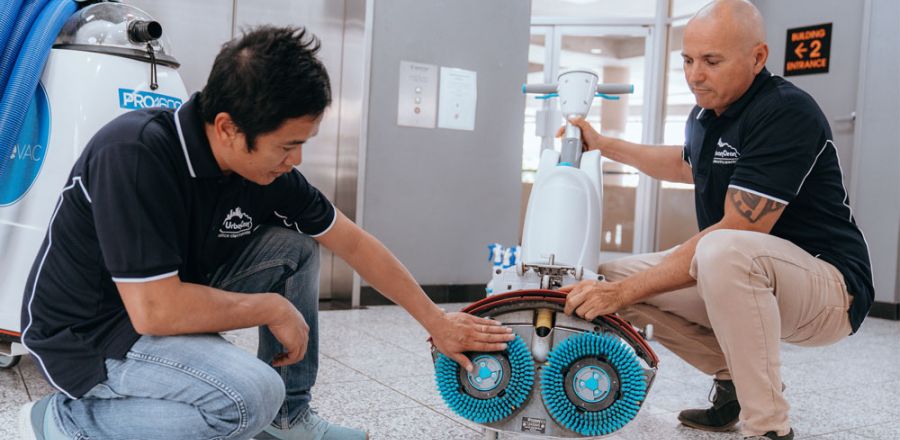Two Urban Clean technicians inspecting a floor-scrubbing machine in an office lobby.